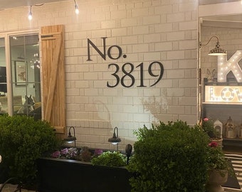 Puede incluir: Una casa de ladrillo blanco con números de casa de metal negro que dicen "No. 3819". Hay luces de cadena colgadas en la parte delantera de la casa y una maceta con arbustos verdes frente a la casa.