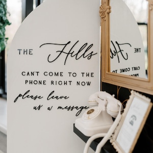 K&ouml;nnte beinhalten: Ein wei&szlig;es, bogenf&ouml;rmiges Schild mit schwarzer Aufschrift THE Hills CAN'T COME TO THE PHONE RIGHT NOW please leave us a message. Ein wei&szlig;es Vintage-W&auml;hlscheibentelefon steht auf einem schwarzen Regal neben einem goldgerahmten Spiegel.