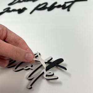Puede incluir: Recortes de nombres en letra cursiva negra con acabado mate. El texto incluye nombres como Daniel, Sarah, James y Robert. Se est&aacute; despegando un trozo de papel protector blanco de la superficie de uno de los recortes de nombres negros.