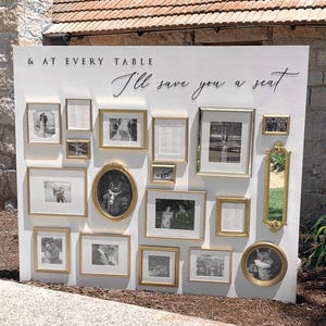 Puede incluir: Un gran panel expositor rectangular blanco con el texto en negro & AT EVERY TABLE I'll save you a seat. El panel est&aacute; decorado con un surtido de marcos dorados de diversas formas y tama&ntilde;os que contienen fotograf&iacute;as en blanco y negro.
