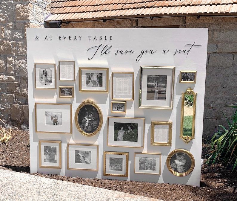 Puede incluir: Un gran panel expositor rectangular blanco con el texto en negro & AT EVERY TABLE I'll save you a seat. El panel est&aacute; decorado con un surtido de marcos dorados de diversas formas y tama&ntilde;os que contienen fotograf&iacute;as en blanco y negro.