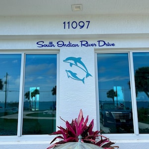 Puede incluir: Un edificio blanco con un letrero azul que dice "11097 South Indian River Drive". Hay dos esculturas de delfines en la pared y una planta roja frente al edificio.
