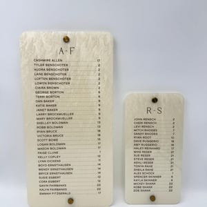 May include: Two rectangular off-white acrylic signs with a marbled texture. The larger sign is labeled A-F and the smaller sign is labeled R-S. Both feature lists of names followed by numbers, secured with small gold-toned circular metal fasteners.