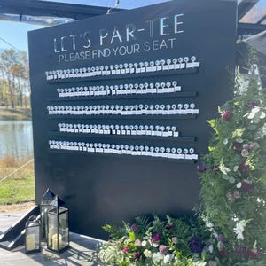 May include: A black seating chart with the text "LET'S PAR-TEE PLEASE FIND YOUR SEAT" in silver lettering. The chart has rows of small white cards attached to it. The scene is decorated with flowers and lanterns.