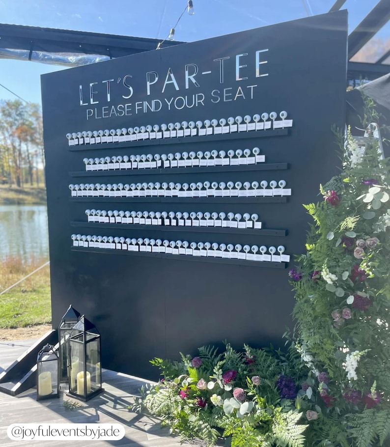 May include: A black seating chart with the text "LET'S PAR-TEE PLEASE FIND YOUR SEAT" in silver lettering. The chart has rows of small white cards attached to it. The scene is decorated with flowers and lanterns.