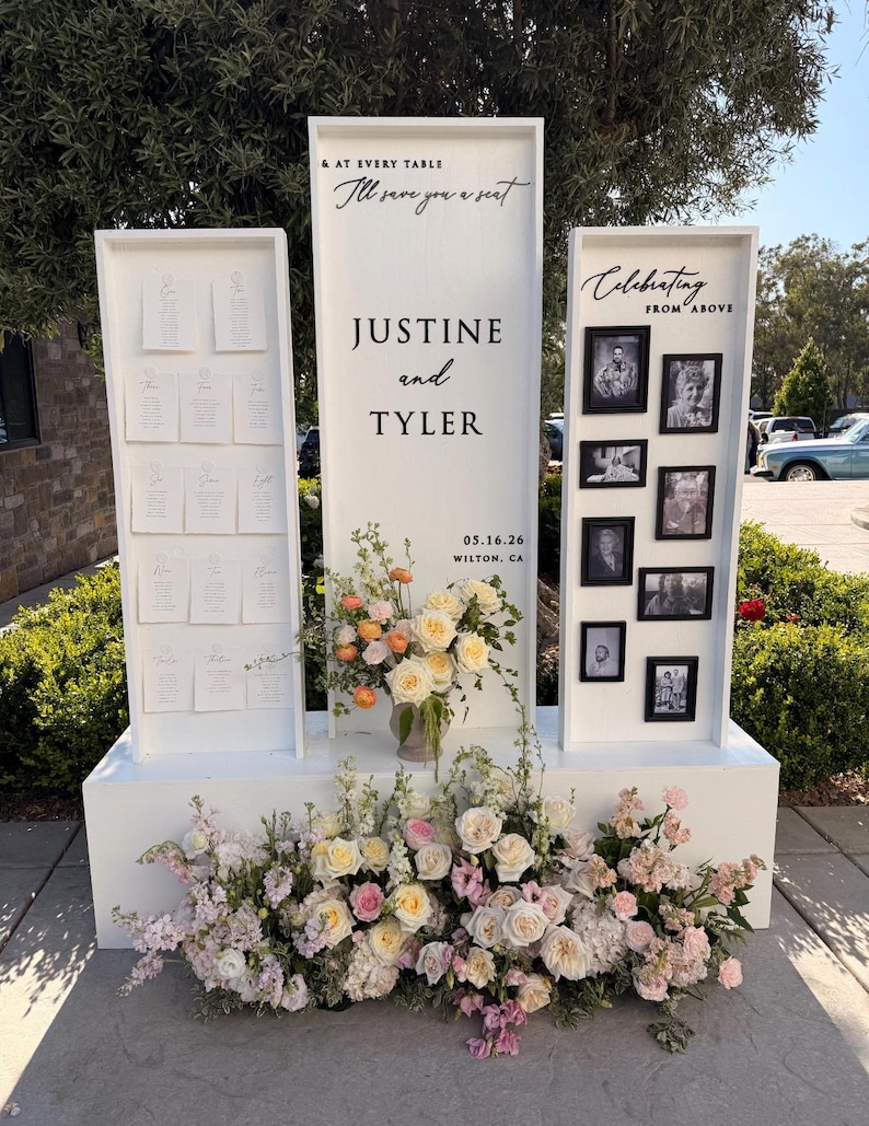 Puede incluir: Un expositor de boda de madera blanca con tres paneles verticales. El panel central dice "& AT EVERY TABLE I'll save you a seat JUSTINE and TYLER 05.16.26 WILTON, CA". El panel izquierdo contiene tarjetas de asiento y el derecho fotos.