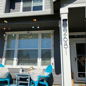 May include: A gray house with a white porch and a blue rug. There are two blue chairs with white cushions and a small wooden table with a sign that says "Good Vibes Only". There are colorful string lights hanging above the porch.