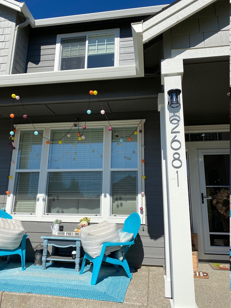 May include: A gray house with a white porch and a blue rug. There are two blue chairs with white cushions and a small wooden table with a sign that says "Good Vibes Only". There are colorful string lights hanging above the porch.
