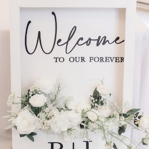 May include: A white framed sign with the words "Welcome to our Forever" in black script. The sign is decorated with white flowers and greenery at the bottom, with the initials "B+L" in black.