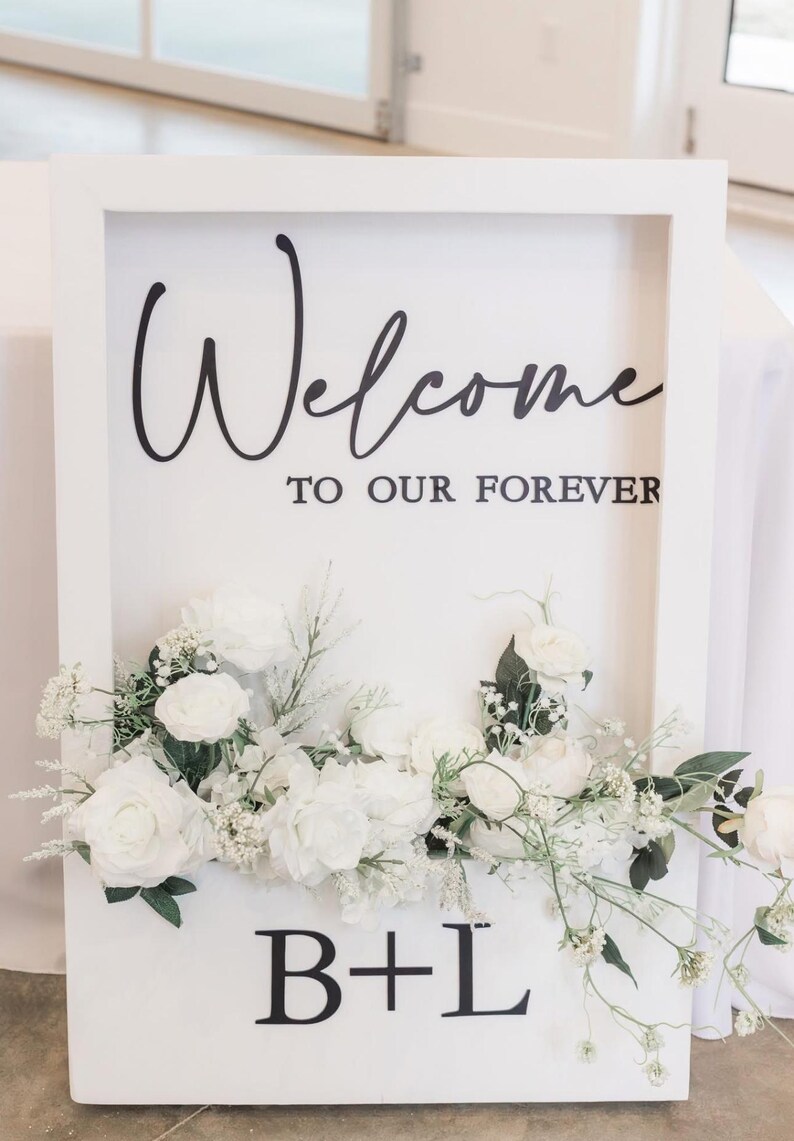 May include: A white framed sign with the words "Welcome to our Forever" in black script. The sign is decorated with white flowers and greenery at the bottom, with the initials "B+L" in black.