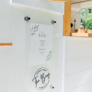 K&ouml;nnte beinhalten: Ein durchsichtiges Acrylschild, das mit schwarzer Hardware an einer wei&szlig;en Paneelwand befestigt ist. Das Schild zeigt ein Men&uuml; mit dem Titel The Bar mit botanischen Blattillustrationen und einem kreisf&ouml;rmigen Logo der Bevy Mobile Bar Company.