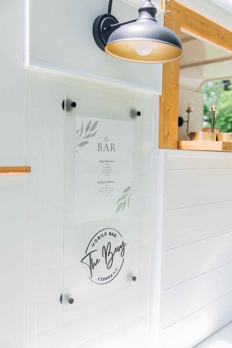 K&ouml;nnte beinhalten: Ein durchsichtiges Acrylschild, das mit schwarzer Hardware an einer wei&szlig;en Paneelwand befestigt ist. Das Schild zeigt ein Men&uuml; mit dem Titel The Bar mit botanischen Blattillustrationen und einem kreisf&ouml;rmigen Logo der Bevy Mobile Bar Company.