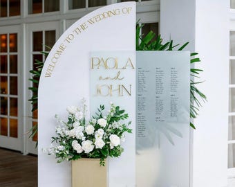 Könnte beinhalten: Hochzeitsschild mit goldener Aufschrift "WELCOME TO THE WEDDING OF PAOLA and JOHN". Es zeigt ein Blumengesteck und einen Sitzplan. Weiße Kerzen in Glashaltern befinden sich am Fuß des Schildes.
