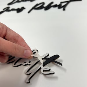 May include: Black and white wooden word cutouts in a cursive font. The word "love" is in the foreground, with protective paper being peeled off. Other names are in the background. The text at the bottom says "Protective paper must be peeled off of front AND back."