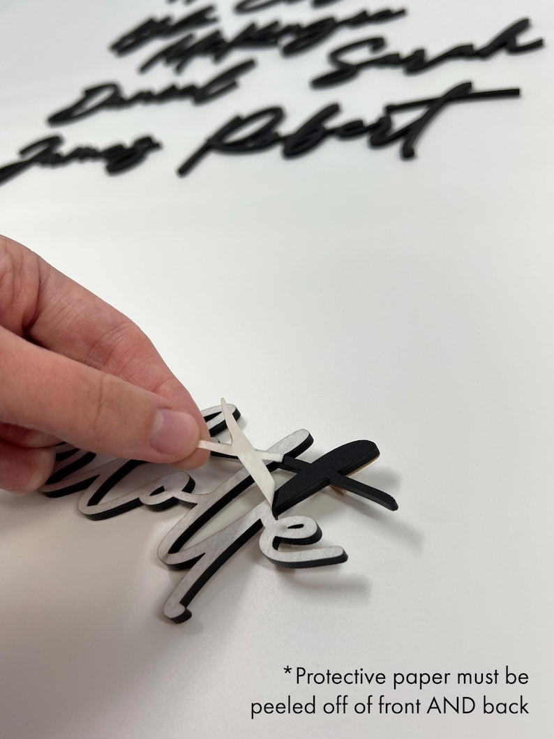 May include: Black and white wooden word cutouts in a cursive font. The word "love" is in the foreground, with protective paper being peeled off. Other names are in the background. The text at the bottom says "Protective paper must be peeled off of front AND back."
