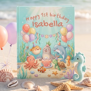 Pode incluir: Um livro de capa dura com uma capa azul-clara apresentando uma cena de aniversário subaquática. O texto diz Happy 1st birthday Isabella. A ilustração inclui uma foca, um golfinho, um polvo, uma tartaruga, um peixe-balão e um cavalo-marinho.