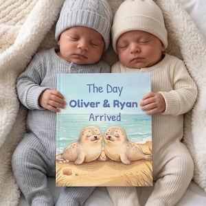 Könnte beinhalten: Ein gebundenes Buch mit dem Titel The Day Oliver & Ryan Arrived, das zwei Robben an einem Sandstrand zeigt. Das Buch wird von zwei Säuglingen in gerippten Langarm-Stramplern und Strickmützen gehalten, die auf einer weichen weißen Decke liegen.