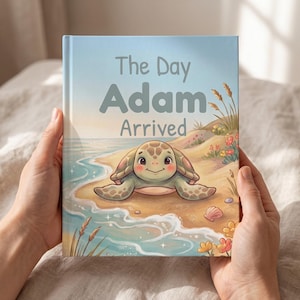 Peut inclure: Un livre à couverture rigide intitulé The Day Adam Arrived présentant une illustration colorée d'une tortue de mer souriante sur une plage de sable avec des vagues, des herbes hautes et des petites fleurs sous un ciel bleu doux.