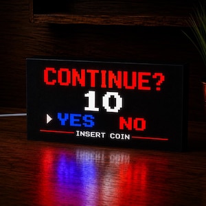 May include: A rectangular black light box featuring a pixelated video game screen display. The text reads CONTINUE? in red, the number 10 in white, YES in blue with a white arrow, NO in red, and INSERT COIN in white at the bottom.