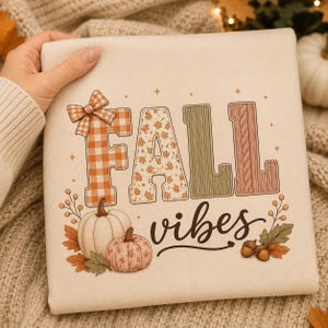 Op de afbeelding: Een crèmekleurig sweatshirt met de tekst FALL in grote blokletters met diverse patronen, waaronder oranje gingham, bloemen, groene ribbels en roze breiwerk. Daaronder staat het woord vibes in cursief schrift met pompoenen.