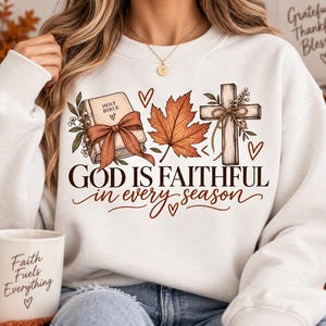 May include: A white crewneck sweatshirt featuring a graphic print of a Holy Bible with a brown ribbon, an orange maple leaf, and a wooden cross. The text reads "GOD IS FAITHFUL in every season" in brown lettering, accompanied by small heart icons.