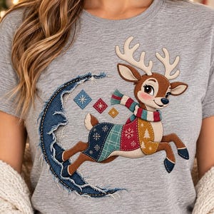 Può includere: Una t-shirt grigio melange con la grafica di una renna marrone che indossa una sciarpa a righe multicolore e un maglione patchwork. La renna salta davanti a una mezzaluna fatta di tessuto denim blu sfilacciato con piccoli rombi.