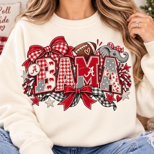 May include: A cream-colored crewneck sweatshirt featuring a graphic print that spells BAMA in large, textured letters. The design includes red and white plaid patterns, a football, a small elephant, red bows, and pom-poms in red and grey.