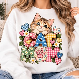 May include: A white crewneck sweatshirt featuring a graphic of a brown tabby kitten peeking over the text CAT MOM. The letters are filled with colorful gingham and floral patterns, surrounded by small hearts, green leaves, and white daisies.