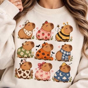 Könnte beinhalten: Ein cremefarbenes Sweatshirt mit einem Grafikdruck von neun Capybaras in verschiedenen Outfits, darunter ein Erdbeerpullover, ein Bienenkostüm, ein Kürbispullover, ein Kuhfleckenmuster, ein blau karierter Schal und ein Pullover mit Mond- und Sternenmuster.