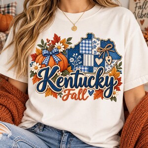 Pode incluir: Uma t-shirt branca com um gráfico de uma abóbora com um laço xadrez azul, um mapa do Kentucky xadrez azul e folhas de outono. O texto Kentucky Fall está escrito em azul e laranja. O design inclui pequenas flores brancas e corações.