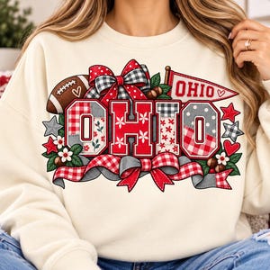 Könnte beinhalten: Ein cremefarbenes Sweatshirt mit Rundhalsausschnitt und einem großen Grafikdruck des Wortes OHIO in Blockbuchstaben. Das Design umfasst rot-weiße Karomuster, einen Football, einen Wimpel, dekorative Schleifen, Sterne, Herzen und Blumen.