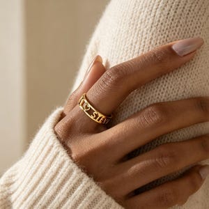 May include: A gold-toned band ring featuring a cut-out design with the name STEVIE and a small heart symbol integrated into the metalwork. The ring has a polished finish and a continuous open-letter style that wraps around the finger.