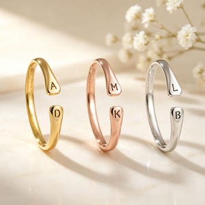 May include: Three open-ended rings in gold, rose gold, and silver tones. Each ring features two flattened, rounded ends, with the letters A and D on the gold ring, M and K on the rose gold ring, and L and B on the silver ring engraved in black.