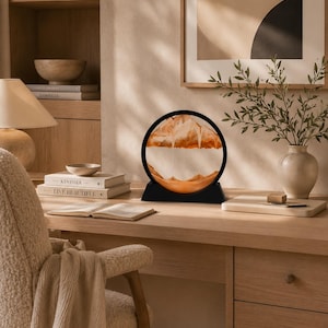 May include: A circular sand art display featuring a black frame and base, containing layers of orange and white sand that form mountain-like shapes as the sand settles within the glass enclosure, creating a dynamic and shifting visual pattern.