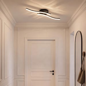Puede incluir: Una lámpara de techo moderna de montaje empotrado con dos brazos metálicos negros ondulados que se cruzan, equipados con tiras de luz LED integradas que emiten un brillo suave y cálido sobre la superficie del techo, creando un efecto luminoso.