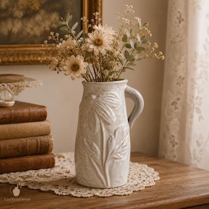 Puede incluir: Un jarrón tipo jarra de cerámica de color crema con un patrón floral en relieve en el exterior. El jarrón tiene una sola asa y está lleno de un ramo de flores secas, que incluye margaritas de colores claros y tallos de follaje verde.