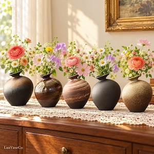 May include: Five round ceramic vases in varying shades of brown, black, and tan, each featuring a narrow neck and filled with a colourful arrangement of fresh flowers including pink ranunculus, white daisies, and purple sweet peas.
