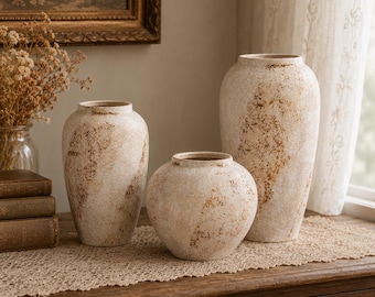 Pode incluir: Três vasos de cerâmica com acabamento texturizado em branco sujo e manchas salpicadas em castanho ferrugem. O conjunto inclui dois vasos altos e esguios e um vaso mais curto e arredondado, todos com uma abertura estreita e aspeto envelhecido.