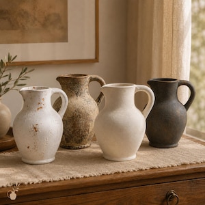 May include: A collection of four ceramic pitchers featuring rounded bodies and side handles. The vessels display varied finishes including distressed white, mottled brown, solid off-white, and dark charcoal, each with a textured, weathered appearance.