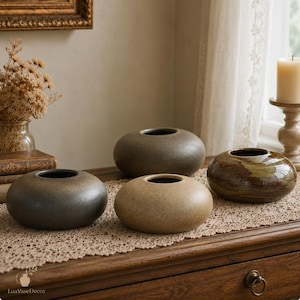 May include: Three low, rounded ceramic vases with smooth, matte and glossy finishes in shades of charcoal grey, beige, and mottled brown. Each vessel features a wide, circular body with a small, centred opening at the top for floral arrangements.