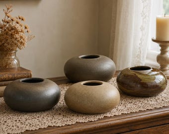 Pode incluir: Três vasos de cerâmica baixos e arredondados com acabamentos lisos, mate e brilhantes em tons de cinza carvão, bege e castanho mosqueado. Cada recipiente apresenta um corpo circular largo com uma pequena abertura centrada no topo.