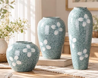 Pode incluir: Três vasos de cerâmica com acabamento salpicado em verde-azulado e branco, cada um decorado com motivos de flores brancas de quatro pétalas. O conjunto inclui um vaso curto arredondado, um oval médio e um alto e esguio, com o logótipo LuaVaseDecor.