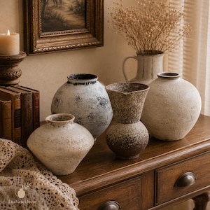 Könnte beinhalten: Eine Sammlung von vier Keramikvasen mit strukturierten Oberflächen. Es gibt eine kleine, hellbeige Vase mit breitem Rand, eine große, runde, abgenutzte weiß-schwarze Vase, eine hohe, gesprenkelte braune Krugvase und eine große, runde, cremeweiße Vase.