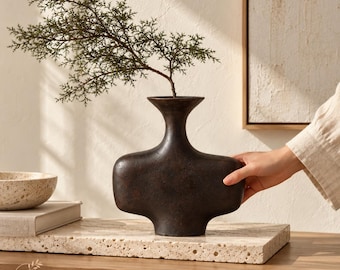 Pode incluir: Um vaso castanho-escuro de corpo largo, com um pescoço estreito e uma borda alargada, apresentando um acabamento texturizado e mate. Um ramo verde com folhas está colocado dentro do vaso. O vaso tem uma forma horizontal distinta com bordas arredondadas.