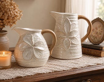 Pode incluir: Duas jarras de cerâmica de cor creme com acabamento salpicado e um design floral em relevo na parte frontal. Cada jarra apresenta uma pega curva lisa de cor castanho claro e uma textura canelada no corpo, com um bico vertedor.