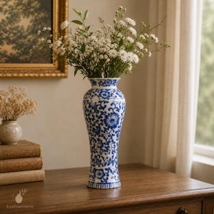 May include: A tall, slender ceramic vase featuring a classic blue and white floral pattern with intricate scrolling vines and blossoms. The vase is filled with a bouquet of small white flowers and green stems, standing upright on a wooden surface.