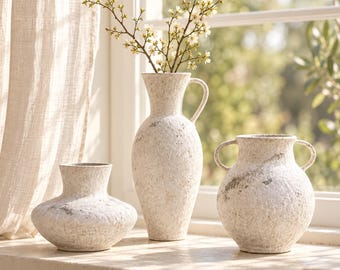 Pode incluir: Três vasos de cerâmica em tom esbranquiçado com acabamento texturizado e desgastado. O conjunto inclui um vaso largo e baixo, um vaso alto tipo jarra com uma alça contendo flores brancas e um vaso redondo com duas alças laterais.