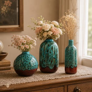 Può includere: Tre vasi in ceramica con finitura smaltata a goccia color turchese e marrone. Il vaso a sinistra è sferico, quello centrale è ovale e quello a destra ha una forma a bottiglia alta. Ognuno contiene composizioni floreali con fiori rosa e bianchi.