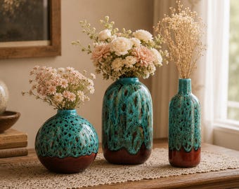 Pode incluir: Três vasos de cerâmica com acabamento em esmalte escorrido turquesa e castanho. O vaso da esquerda é esférico, o central é oval e o da direita tem forma de garrafa alta. Cada um contém arranjos florais com flores rosa claro e brancas.
