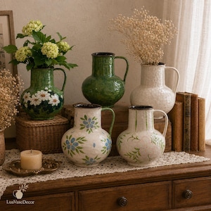 Op de afbeelding: Vijf keramische kannen met oren, voorzien van diverse ontwerpen waaronder effen groen, wit met blauwe bloemen, wit met groen blad en groen met witte madeliefjes. De kannen hebben een getextureerde afwerking en donkere randen.